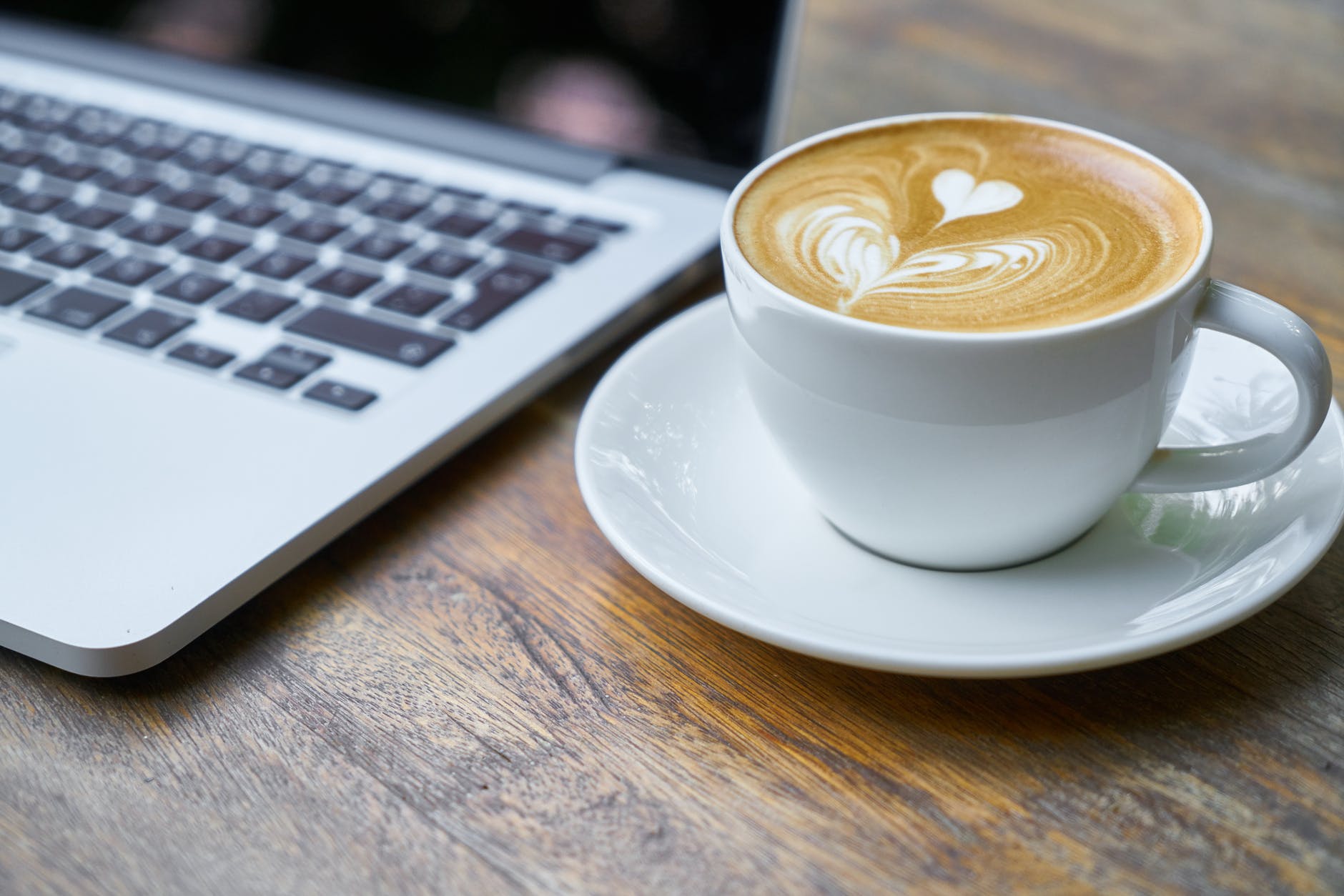 Fancy coffee in a cup and saucer on a wooden table next to a laptop.